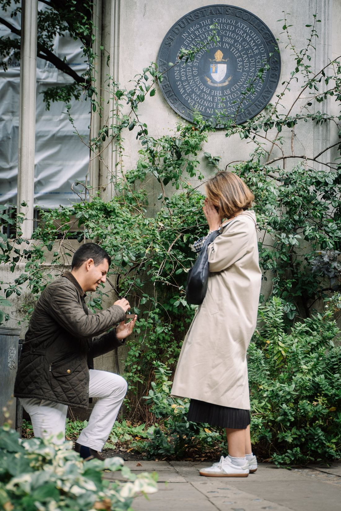 Man proposing by an ivy wall as she covers her face