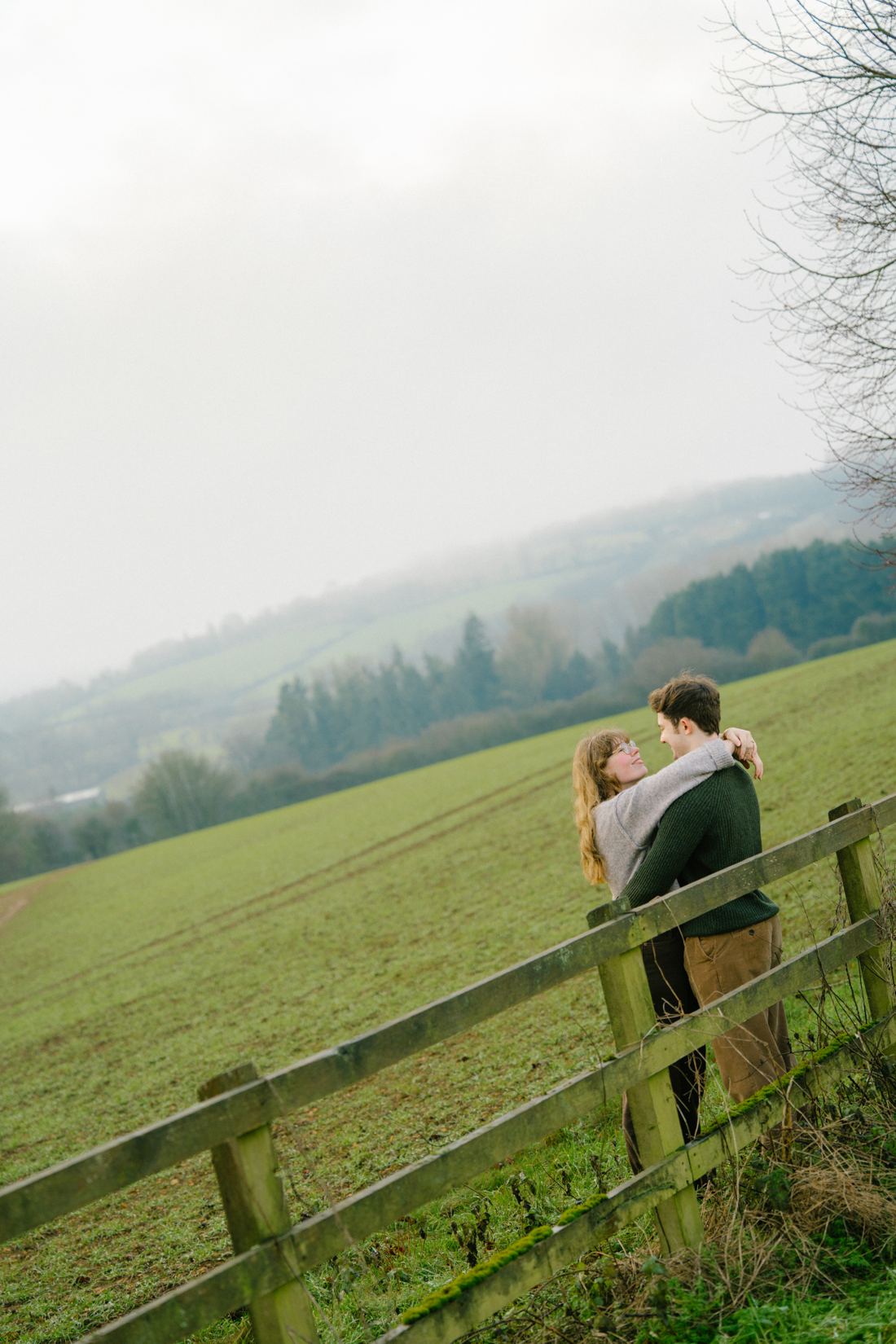 Couple hugging by a wooden fence on a misty Cotswolds hillside